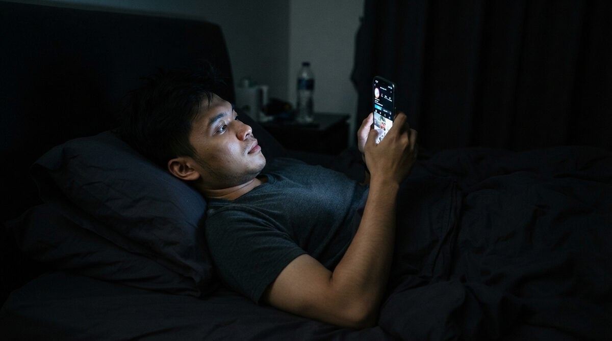 A person lying in bed in a dark room late at night, illuminated only by the glowing screen of a smartphone.